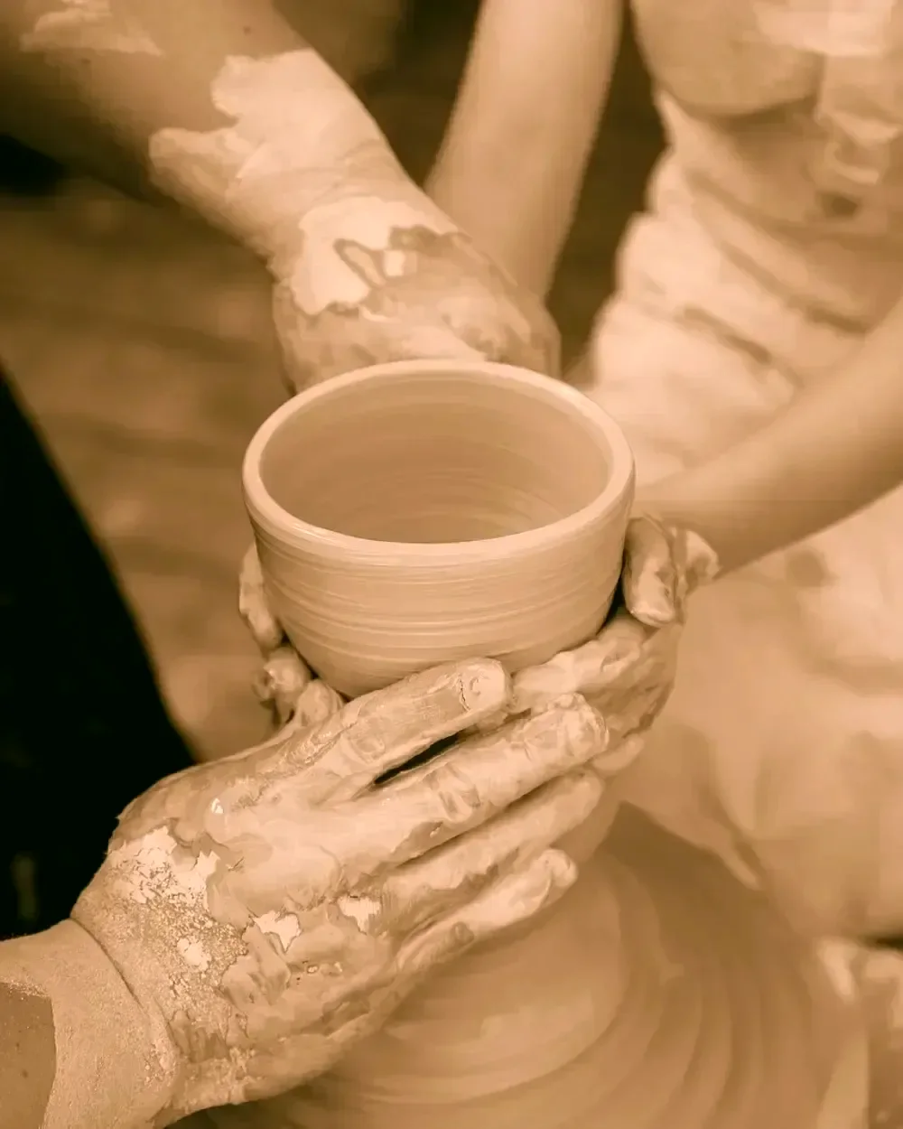 Two pairs of clay-covered hands cradling a freshly thrown bowl still spinning on the wheel.